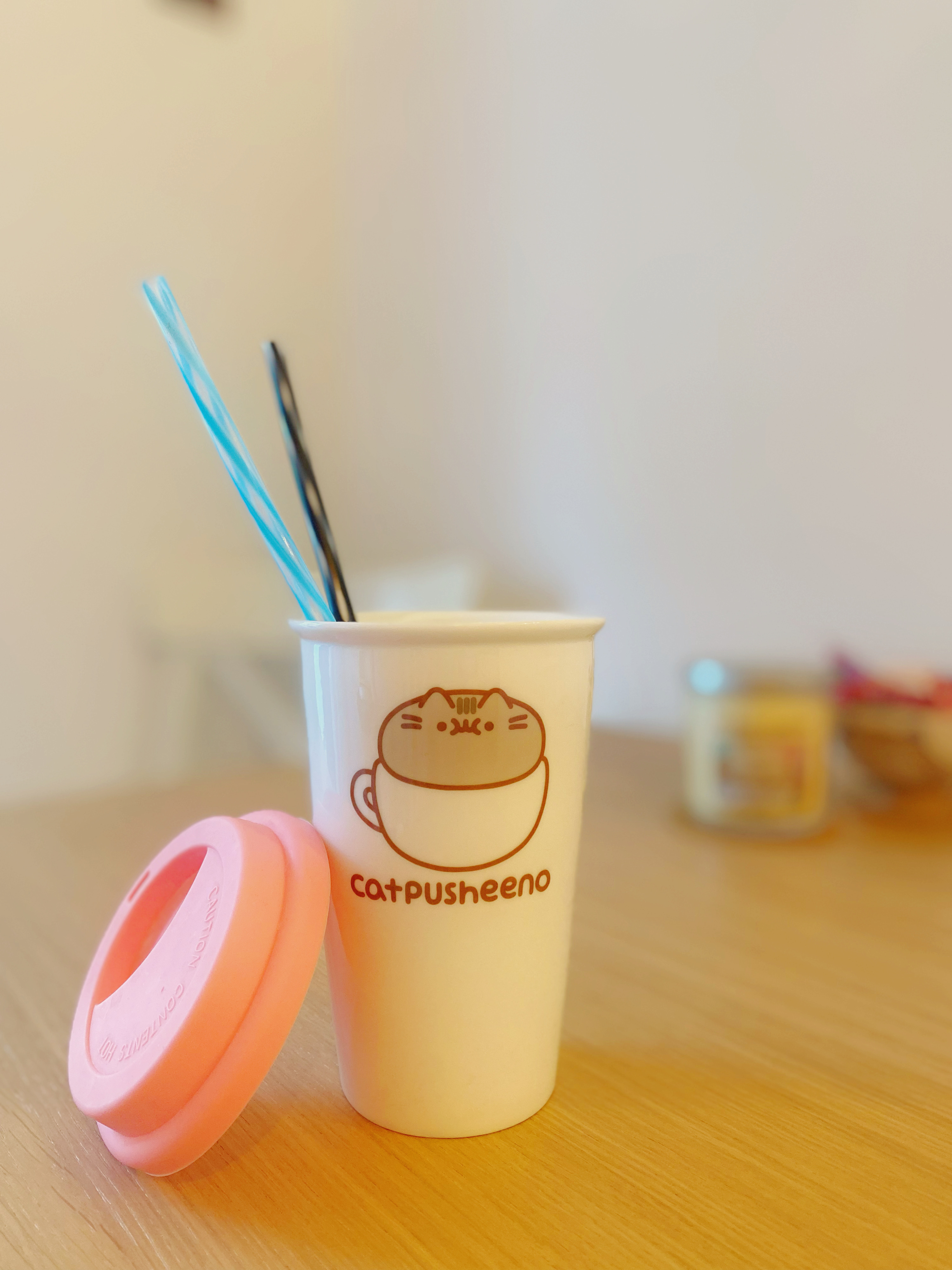 Becher mit Deckel und Trinkhalm mit einem Pusheen-Cat-Design, auf einem Holzuntergrund.