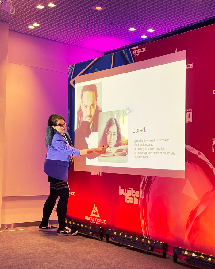 Frau in blauer Jacke hält eine Präsentation auf einer Bühne bei der TwitchCon, zeigt auf eine Folie mit dem Titel &apos;Bored&apos;.