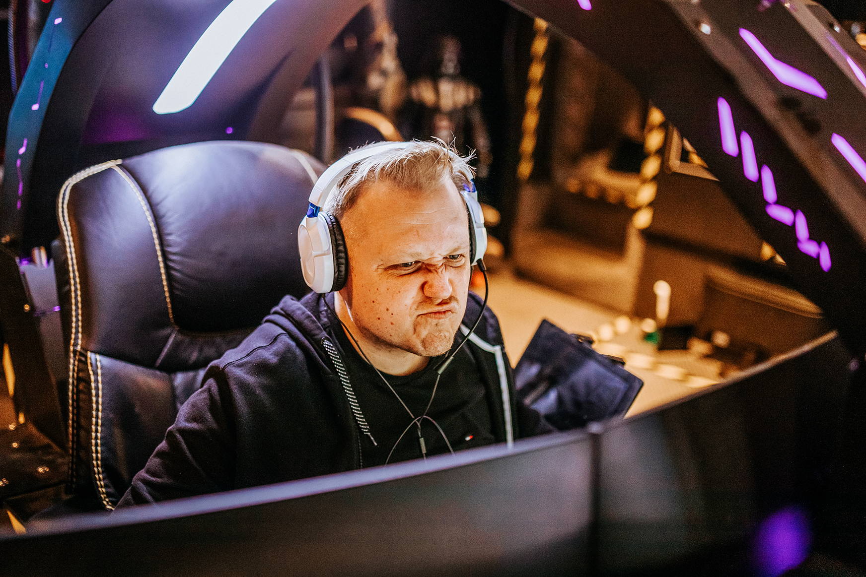 Mann in einem Gaming-Stuhl mit Headset, konzentriert auf seinen Bildschirm, umgeben von Beleuchtung in einem futuristischen Raum.