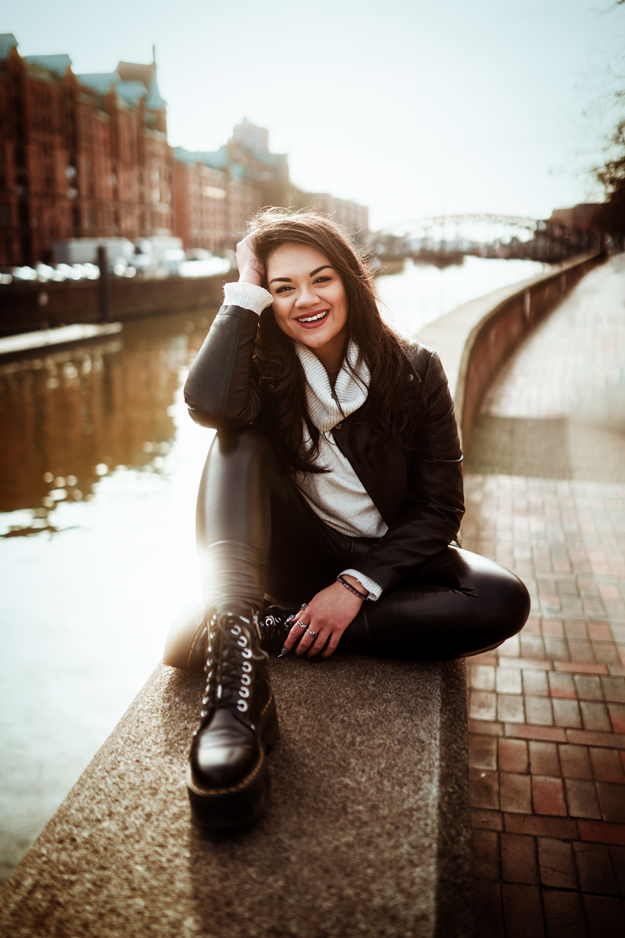 Lächelnde Frau in schwarzer Lederjacke und Lederleggings sitzt auf einer Steinmauer an einem Kanal, umgeben von sonnigem Wetter und städtischer Architektur, bekannt als Sabine Gasparini.