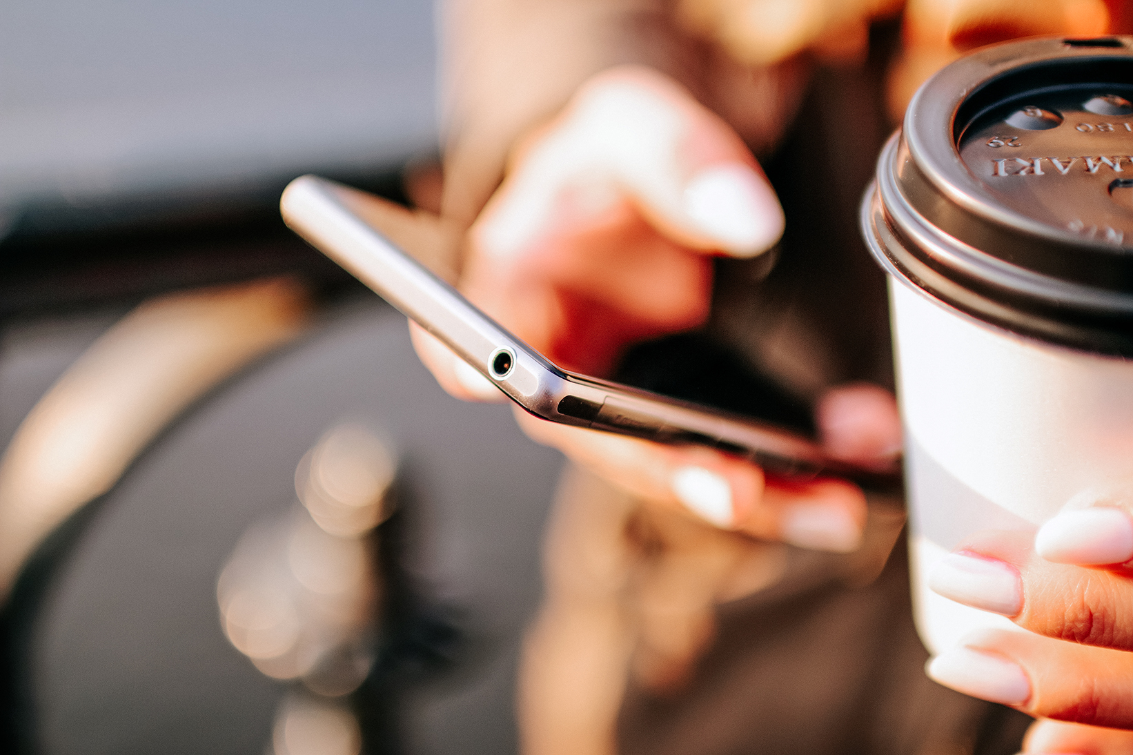 Person hält ein Smartphone und einen Kaffeebecher, beide Hände sind scharf im Fokus.