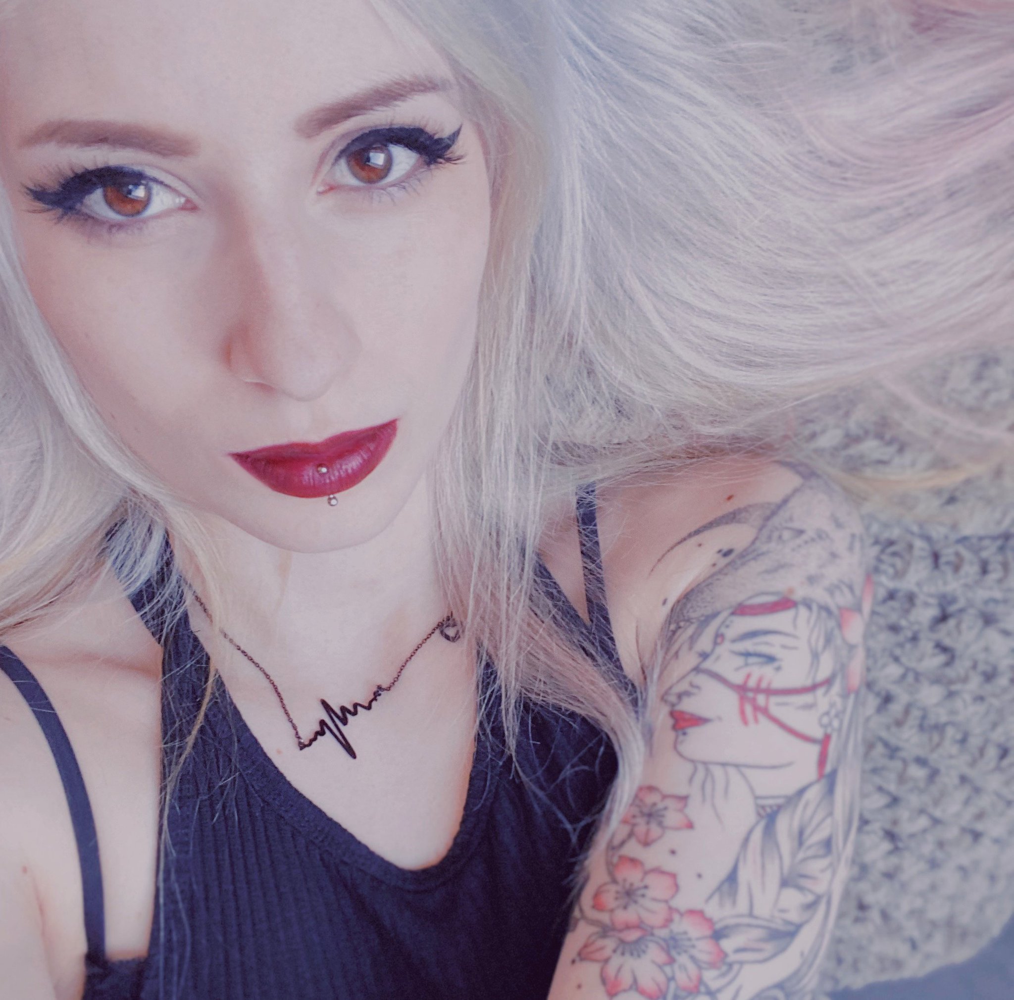 Junge Frau mit langen blonden Haaren, rotem Lippenstift und schwarzem Oberteil, trägt eine Halskette mit Herzschlag-Symbol und zeigt Tätowierungen am Arm, bekannt als JustTrashee.