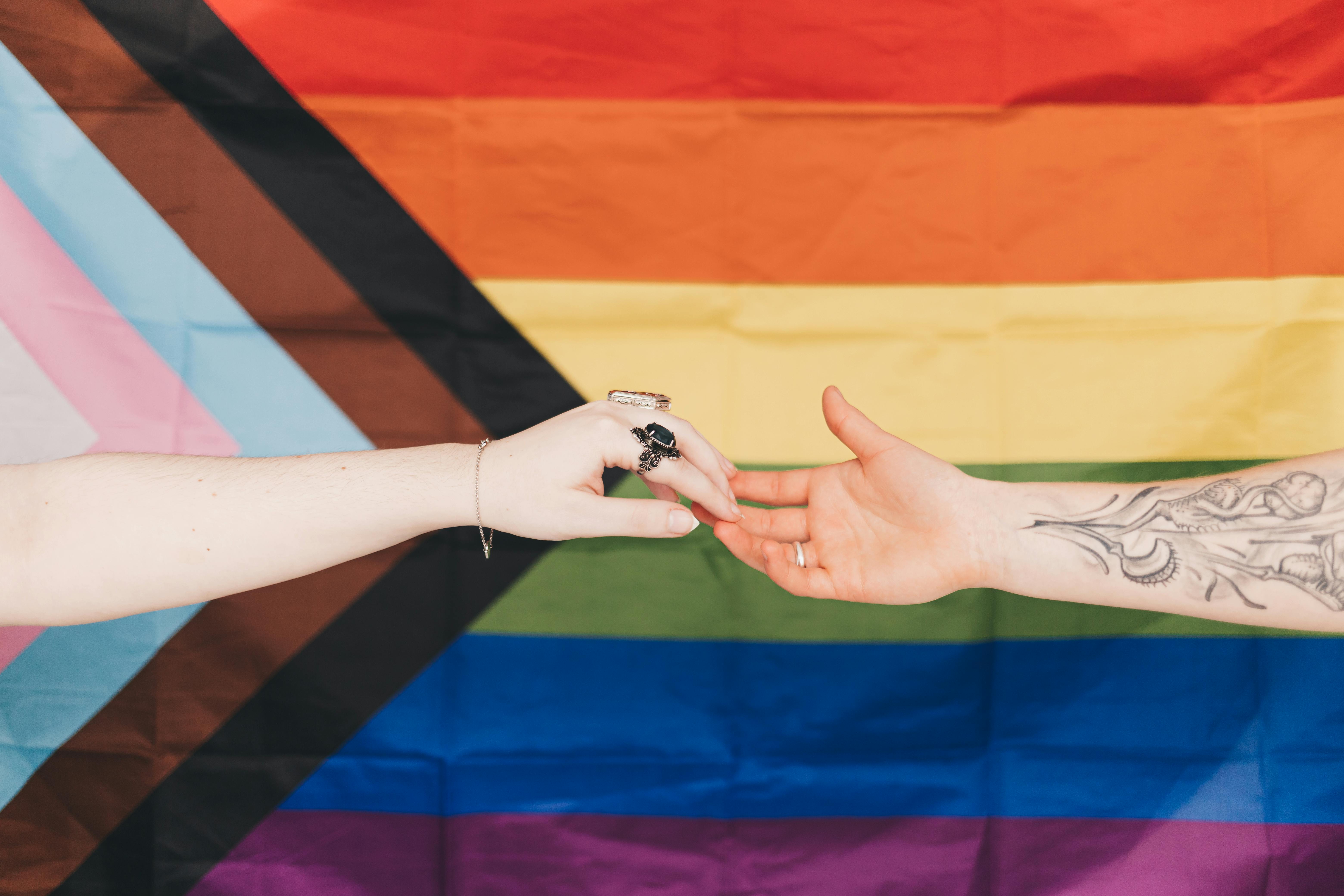 Zwei Hände berühren sich vor einer Progress-Pride-Flagge, symbolisieren Unterstützung und Zusammenhalt.