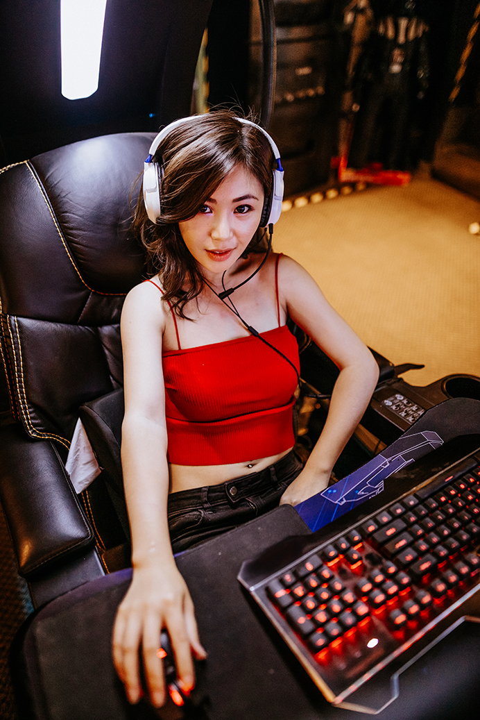 Junge Frau mit Headset in einem Gaming-Stuhl, trägt ein rotes Top und sitzt vor einer beleuchteten Tastatur, bereit zum Spielen.
